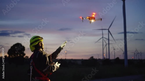Two Asian technicians in a safety kit fly the drone to inspect the wind turbine's operation.