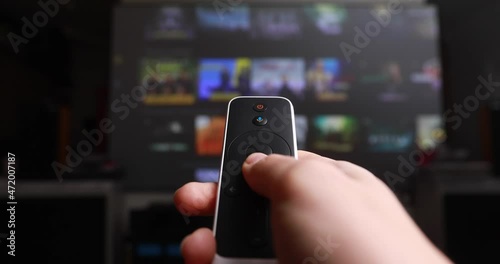 Womans hand selects internet tv channels with remote control. Close up view. Blurry tv at the background
