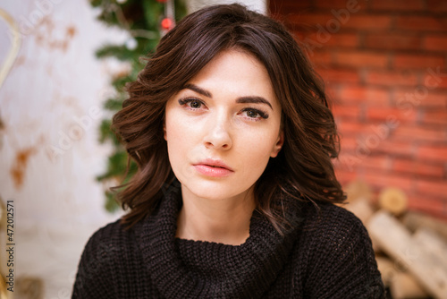 Close-up portrait of woman looking at camera against background of fireplace with christmas decor. Beautiful young brunette caucasian appearance in a black sweater gazes intently with brown eyes