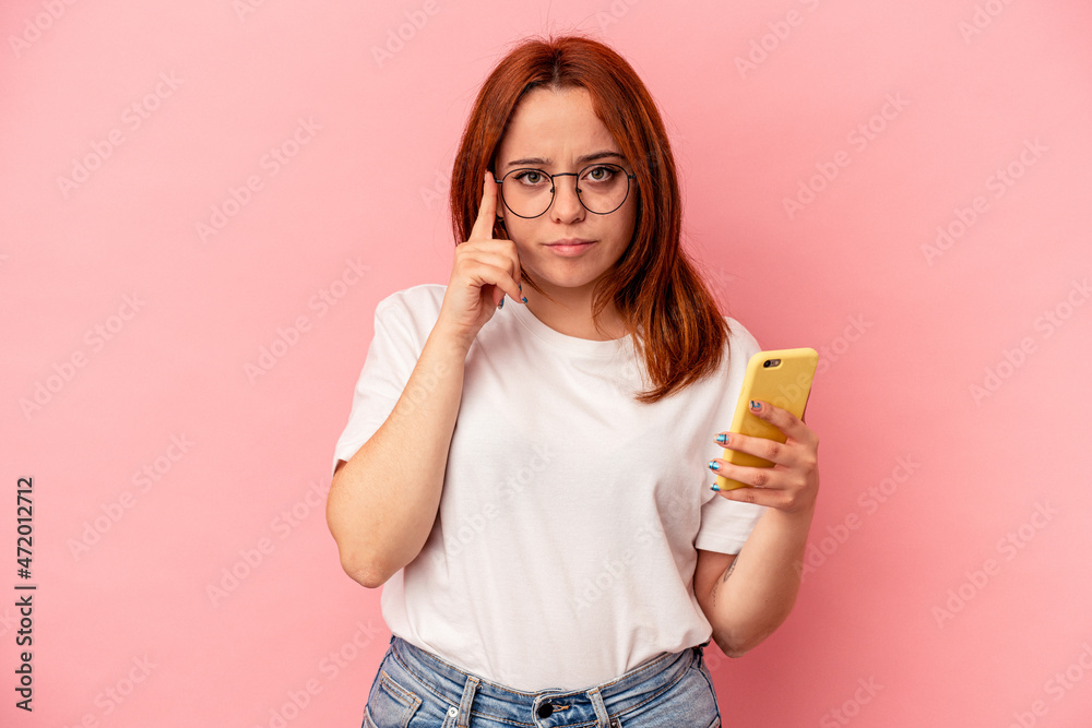 Young caucasian woman holding a mobile phone isolated on pink background pointing temple with finger, thinking, focused on a task. Young caucasian woman holding a mobile phone isolated on pink background pointing temple with finger, thinking, focused on a task.