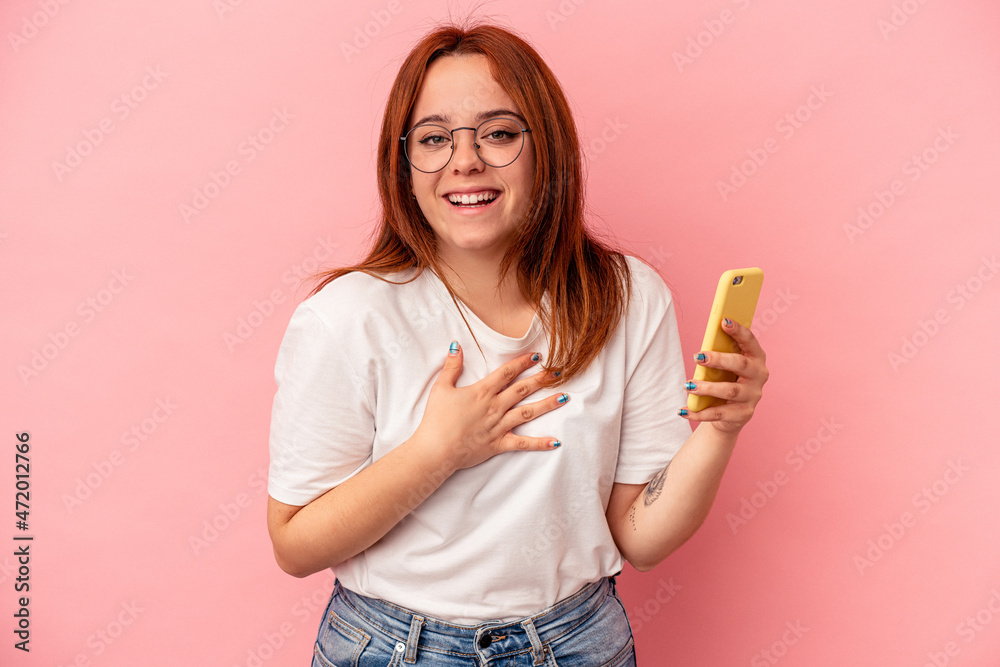 Young caucasian woman holding a mobile phone isolated on pink background laughs out loudly keeping hand on chest. Young caucasian woman holding a mobile phone isolated on pink background laughs out loudly keeping hand on chest.