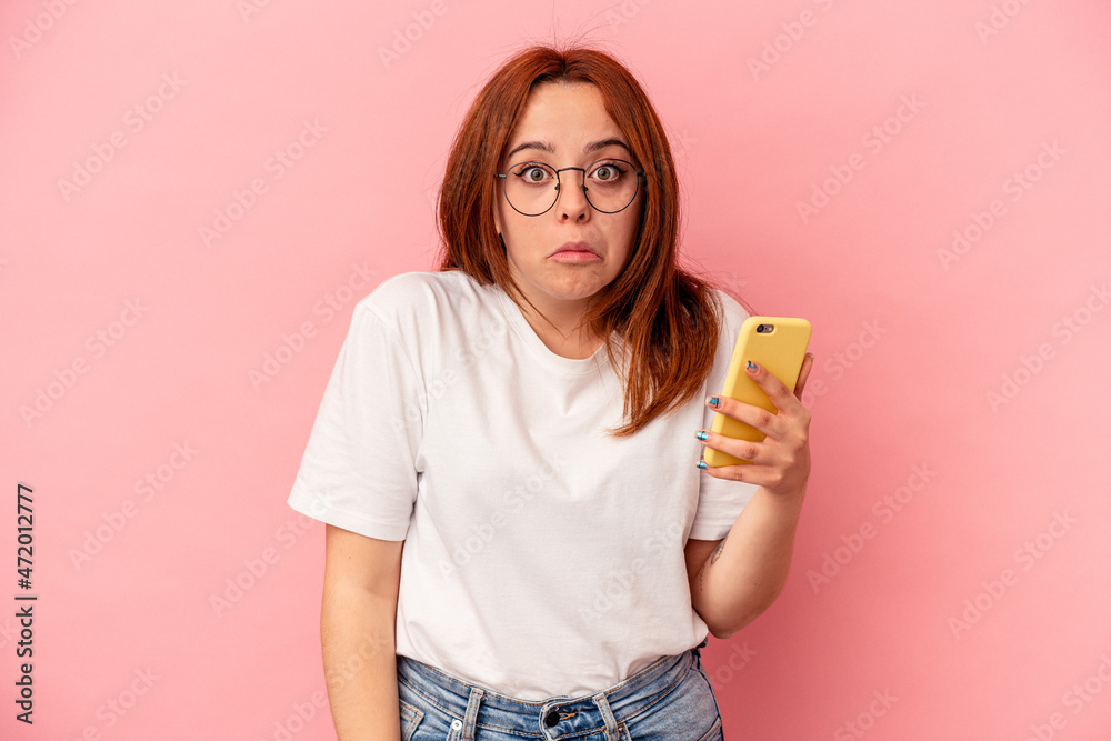 Young caucasian woman holding a mobile phone isolated on pink background shrugs shoulders and open eyes confused. Young caucasian woman holding a mobile phone isolated on pink background shrugs shoulders and open eyes confused.