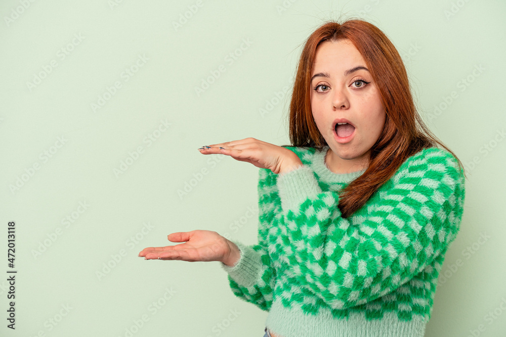 Young caucasian woman isolated on green background shocked and amazed holding a copy space between hands. Young caucasian woman isolated on green background shocked and amazed holding a copy space between hands.