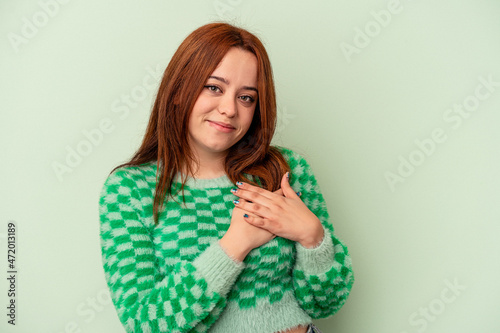 Young caucasian woman isolated on green background has friendly expression, pressing palm to chest. Love concept. © Asier