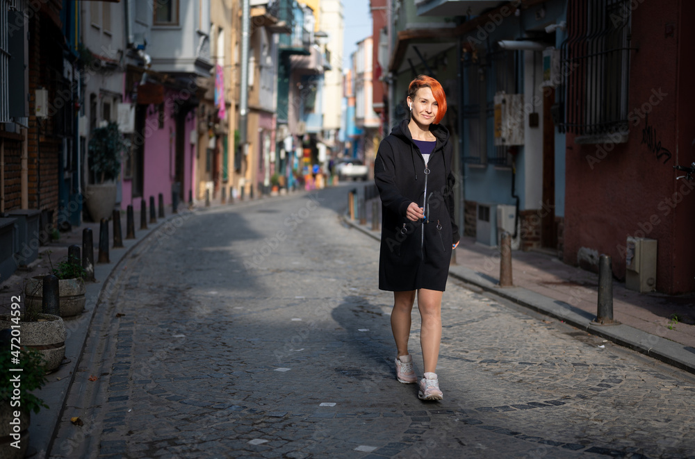 Young lesbian woman confidently walks down street and looks ahead. Young cheerful woman with bright red hair walks down a narrow street