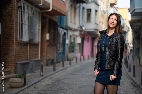 Tall attractive woman walks along a narrow city street. Young woman is wearing a leather jacket and short dress. look into the distance © Celt Studio