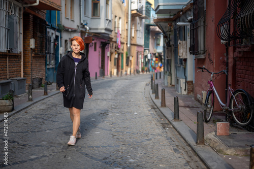 Young lesbian woman confidently walks down the street and looks ahead. Young cheerful woman with bright red hair walks down a narrow street