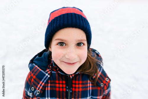 Fotografi Close up funny portrait of little caucasian girl in winter clothes looking at camera and smiling, standing on snow field