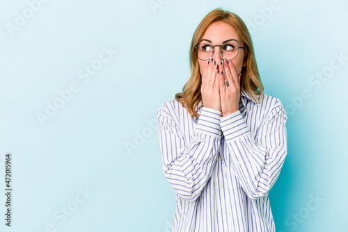 Young caucasian woman isolated on blue background thoughtful looking to a copy space covering mouth with hand.