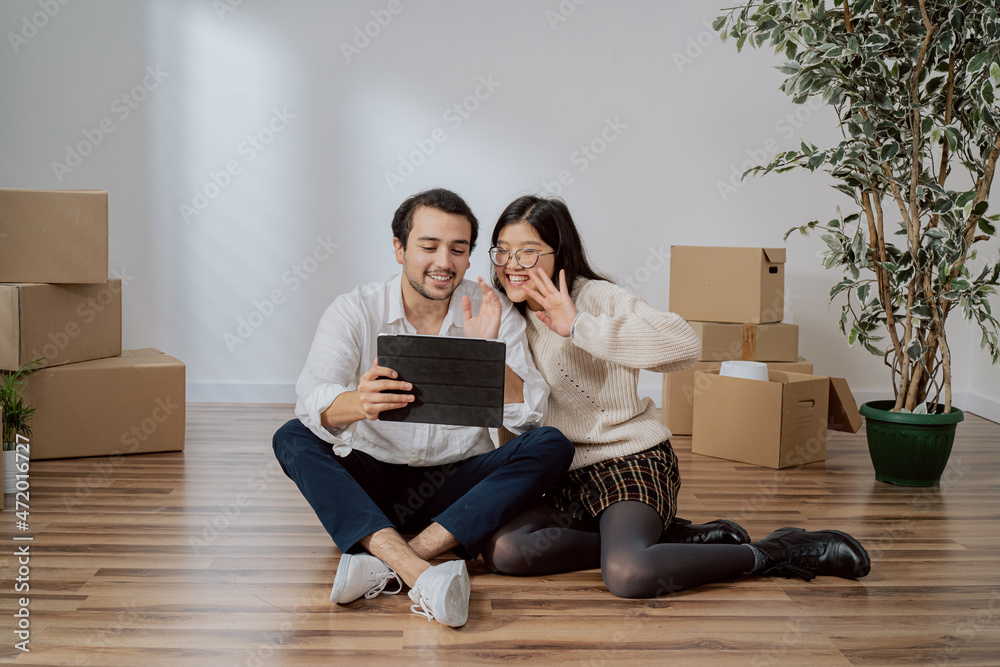 Lovers are sitting on floor in newly bought apartment elegantly dressed man is holding tablet couple is chatting with parents friends over internet waving to camera bragging about moving into flat Lovers are sitting on floor in newly bought apartment elegantly dressed man is holding tablet couple is chatting with parents friends over internet waving to camera bragging about moving into flat