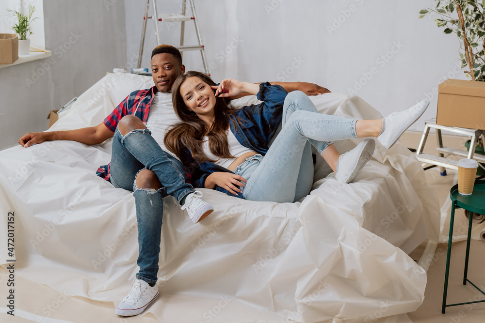 Smiling couple in love, married of different nationalities lie on couch protected by foil, interior of the apartment during renovation, rearrangement, painting walls, changing the color of the room Smiling couple in love, married of different nationalities lie on couch protected by foil, interior of the apartment during renovation, rearrangement, painting walls, changing the color of the room