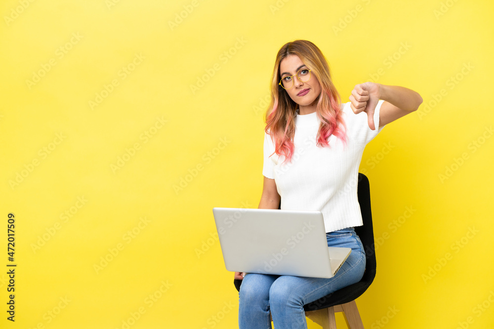 Young woman sitting on a chair with laptop over isolated yellow background showing thumb down with negative expression Young woman sitting on a chair with laptop over isolated yellow background showing thumb down with negative expression