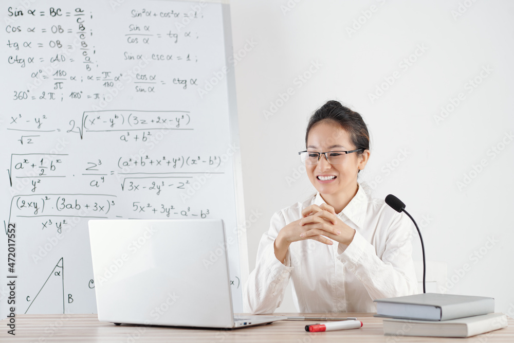 © DragonImages - Smiling young college teacher answering questions of students during online class © DragonImages - Smiling young college teacher answering questions of students during online class