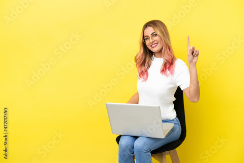 Young woman sitting on a chair with laptop over isolated yellow background showing and lifting a finger in sign of the best