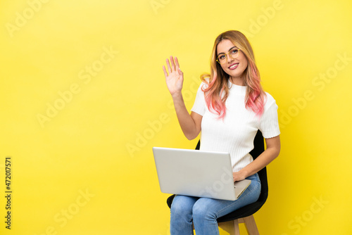 Young woman sitting on a chair with laptop over isolated yellow background saluting with hand with happy expression © luismolinero