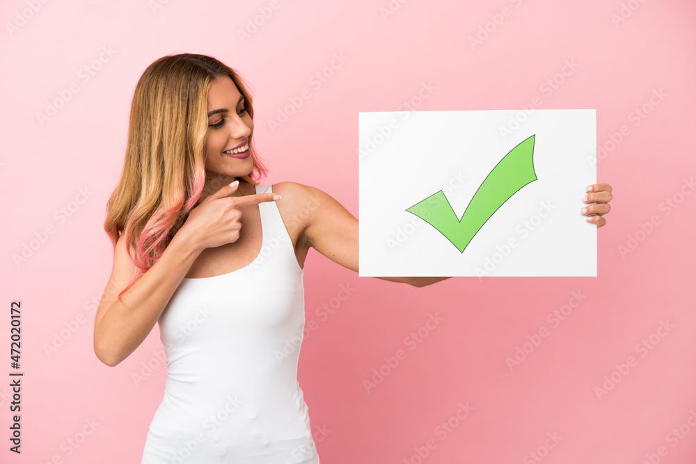 Young woman over isolated pink background holding a placard with text Green check mark icon and pointing it Young woman over isolated pink background holding a placard with text Green check mark icon and pointing it