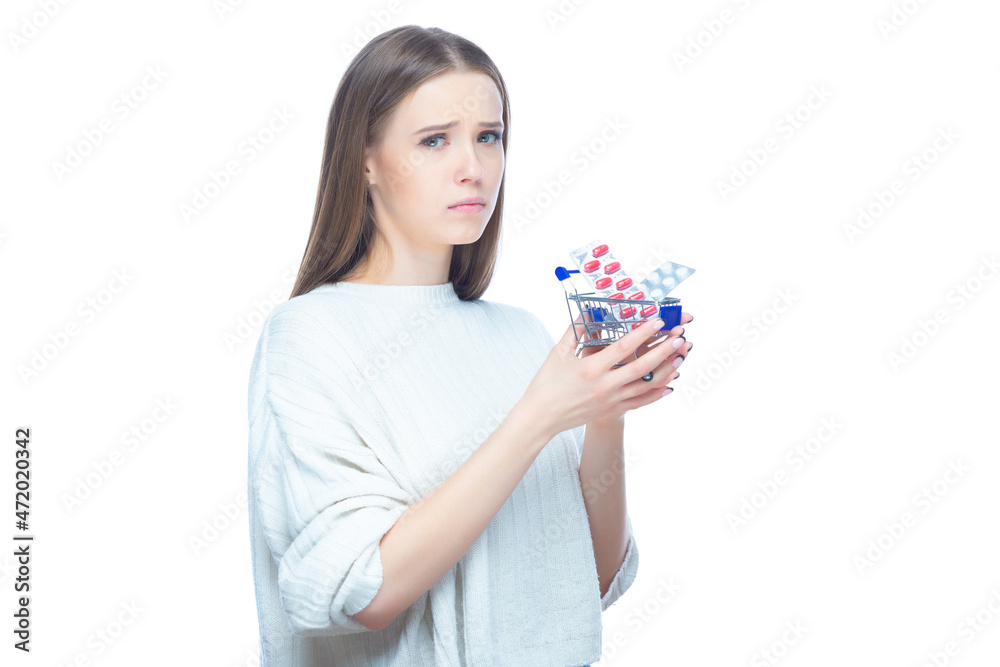 Studio shot of upset disappointed girl holding in her hands a small shopping cart full of expensive pills and medicine, isolated on white background Studio shot of upset disappointed girl holding in her hands a small shopping cart full of expensive pills and medicine, isolated on white background