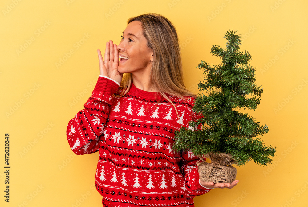 Young caucasian woman holding a little christmas tree isolated on yellow background shouting and holding palm near opened mouth. Young caucasian woman holding a little christmas tree isolated on yellow background shouting and holding palm near opened mouth.