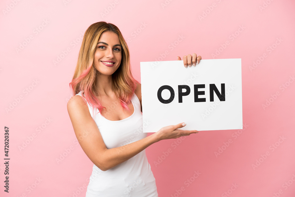Young woman over isolated pink background holding a placard with text OPEN with happy expression Young woman over isolated pink background holding a placard with text OPEN with happy expression