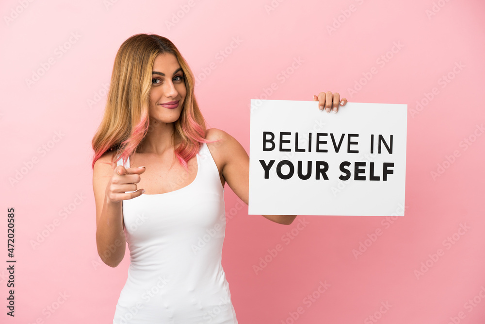 Young woman over isolated pink background holding a placard with text Believe In Your Self and pointing to the front Young woman over isolated pink background holding a placard with text Believe In Your Self and pointing to the front
