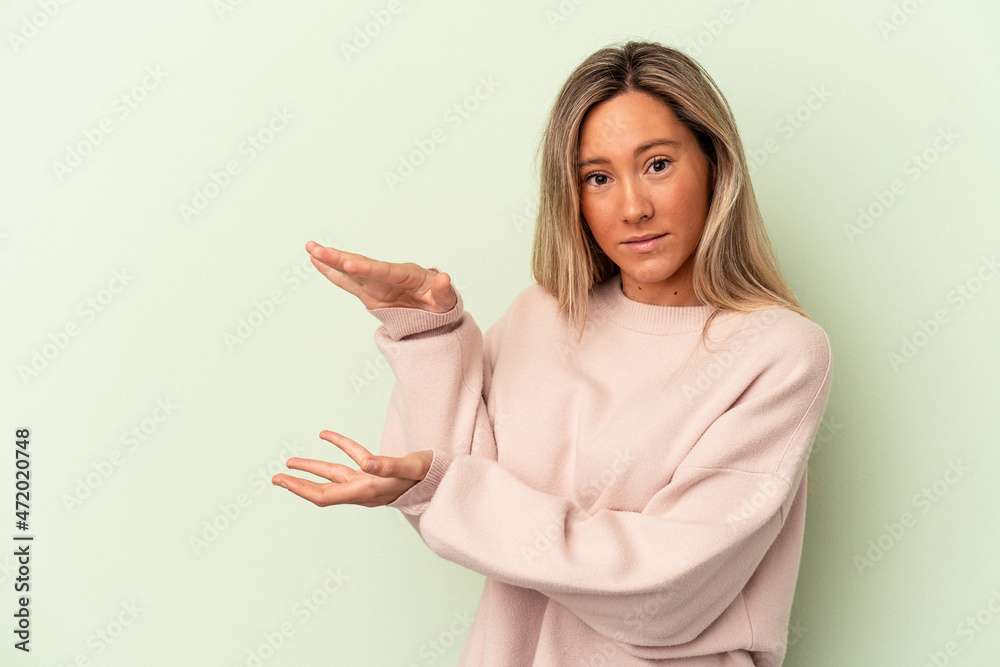 Young caucasian woman isolated on green background shocked and amazed holding a copy space between hands. Young caucasian woman isolated on green background shocked and amazed holding a copy space between hands.