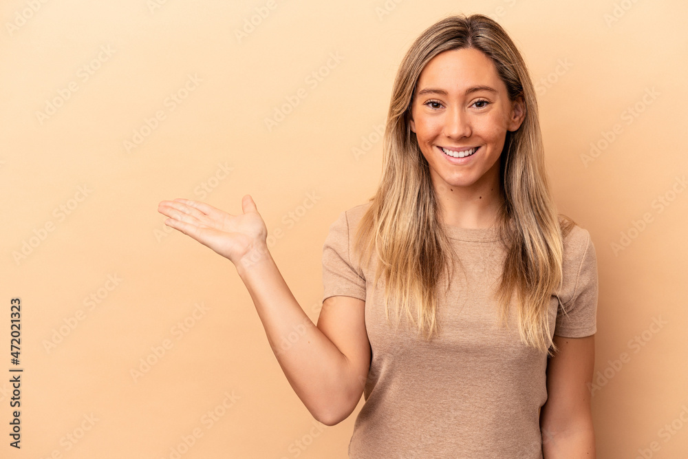 Young caucasian woman isolated on beige background showing a copy space on a palm and holding another hand on waist. Young caucasian woman isolated on beige background showing a copy space on a palm and holding another hand on waist.