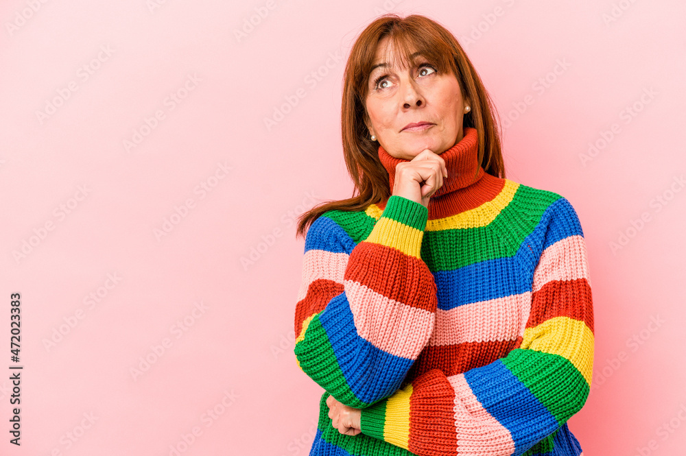 Middle age caucasian woman wearing a multicolor sweater isolated on pink background looking sideways with doubtful and skeptical expression.