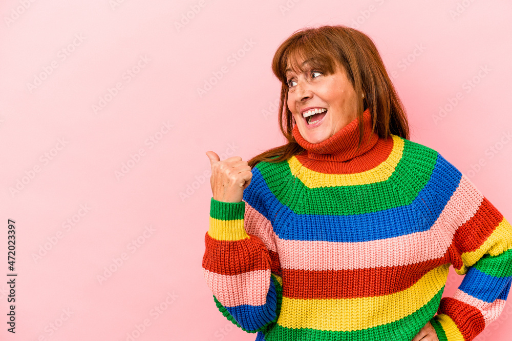 Middle age caucasian woman wearing a multicolor sweater isolated on pink background points with thumb finger away, laughing and carefree. Middle age caucasian woman wearing a multicolor sweater isolated on pink background points with thumb finger away, laughing and carefree.