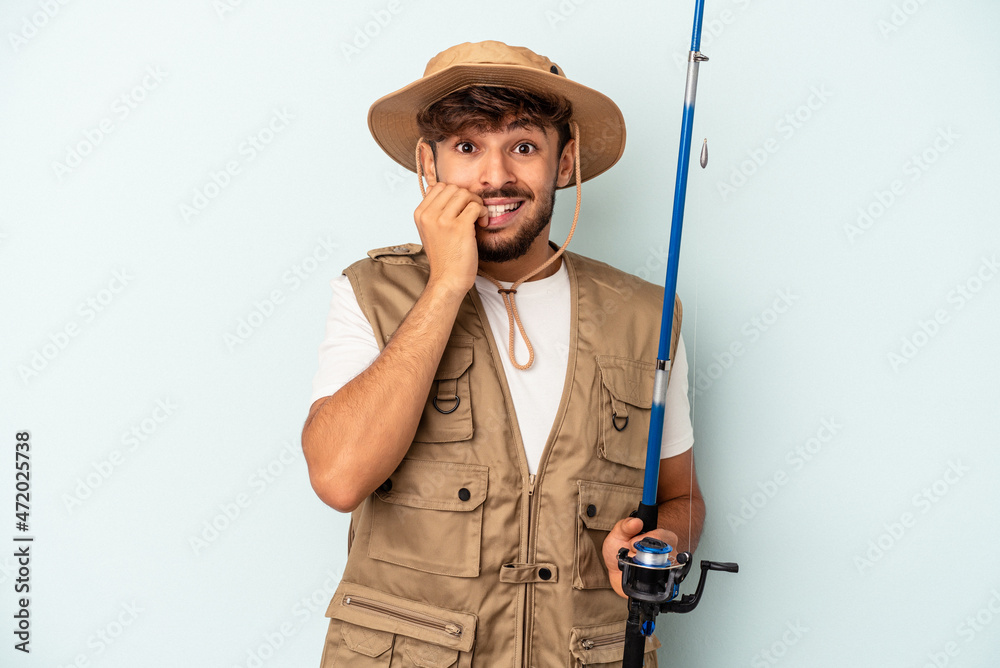 Young mixed race fisherman holding a rod isolated on blue background biting fingernails, nervous and very anxious. Young mixed race fisherman holding a rod isolated on blue background biting fingernails, nervous and very anxious.