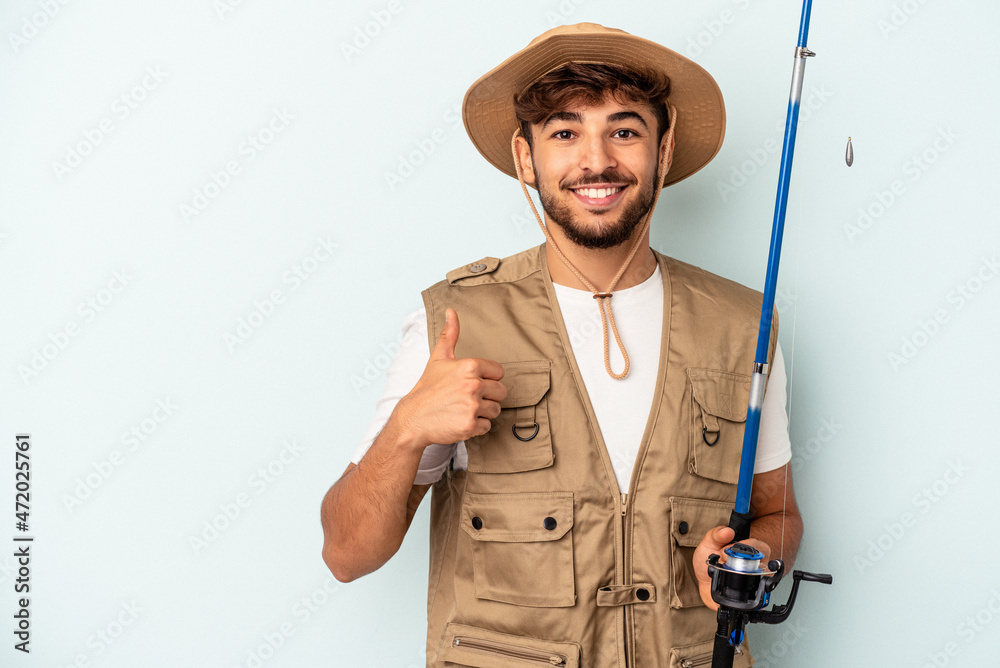 Young mixed race fisherman holding a rod isolated on blue background smiling and raising thumb up