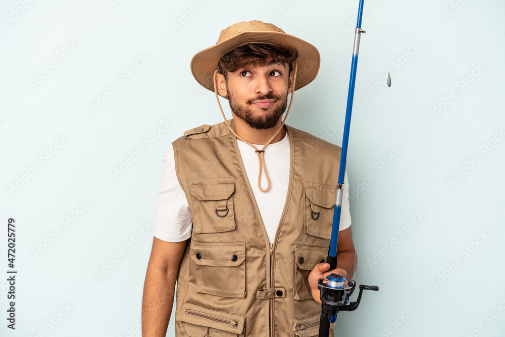 Young mixed race fisherman holding a rod isolated on blue background confused, feels doubtful and unsure. Young mixed race fisherman holding a rod isolated on blue background confused, feels doubtful and unsure.