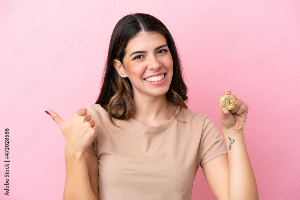 Young Italian woman holding a Bitcoin isolated on pink background pointing to the side to present a product Young Italian woman holding a Bitcoin isolated on pink background pointing to the side to present a product