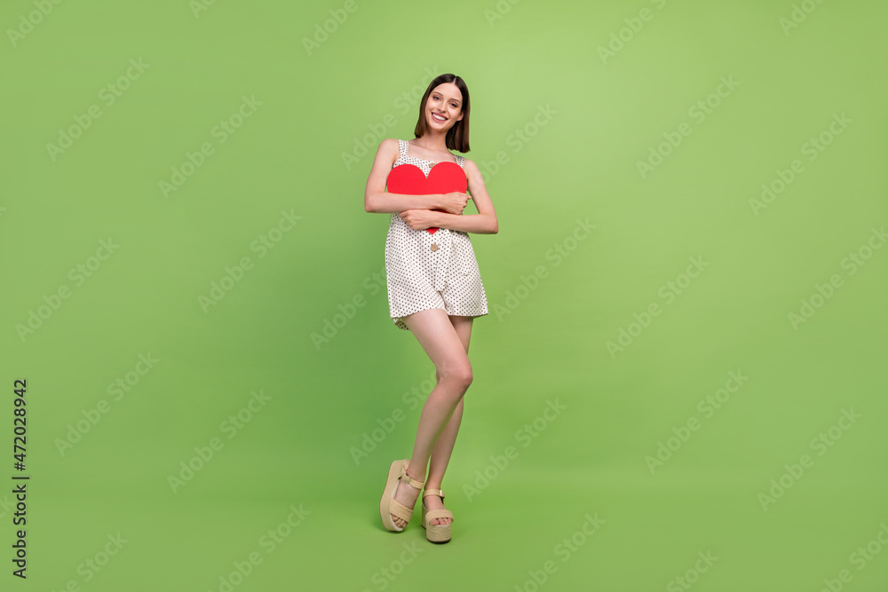 Full body photo of cute millennial brunette lady hold heart wear overall sandals isolated on green background Full body photo of cute millennial brunette lady hold heart wear overall sandals isolated on green background