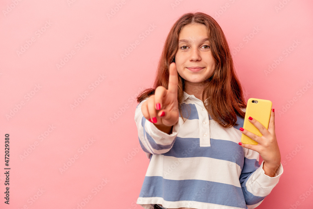 Little caucasian girl holding mobile phone isolated on pink background showing number one with finger. Little caucasian girl holding mobile phone isolated on pink background showing number one with finger.