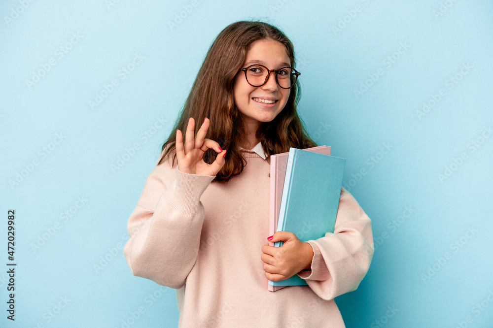 Little caucasian student girl holding books isolated on blue background cheerful and confident showing ok gesture. Little caucasian student girl holding books isolated on blue background cheerful and confident showing ok gesture.