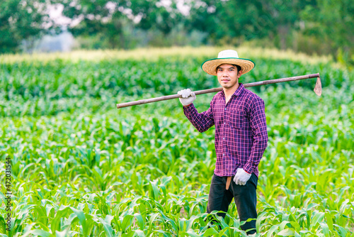 Man Asian farmer wearing a red plaid shirt and white gloves carry a hoe on shoulder in green corn field .