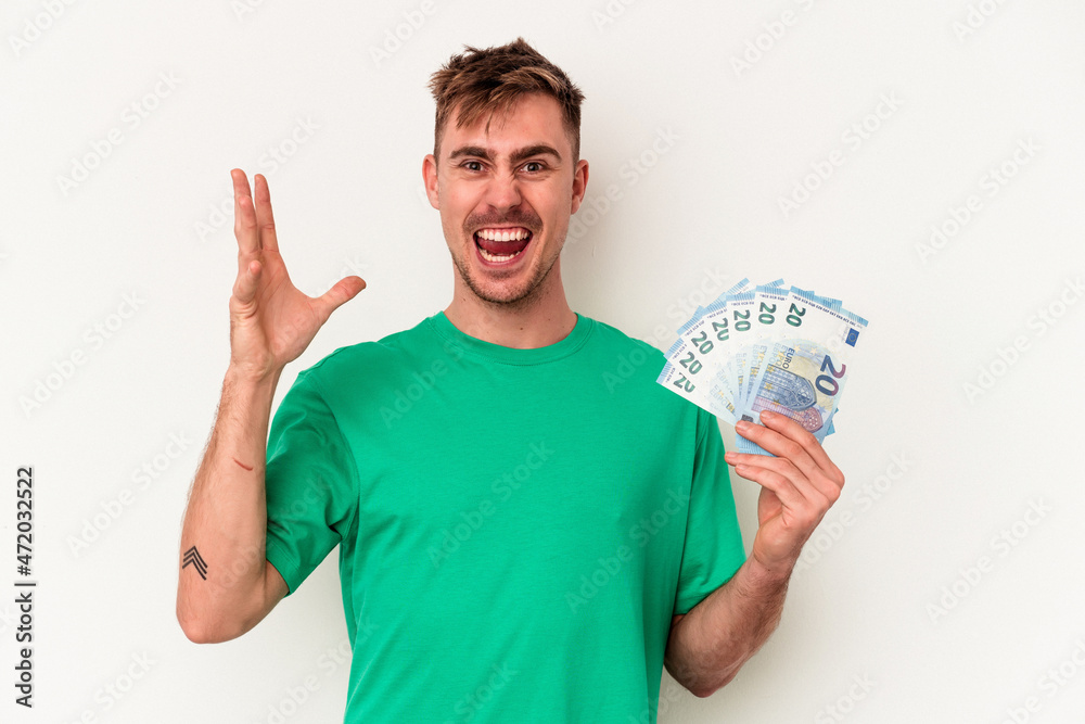 Young caucasian man holding bank notes isolated on white background receiving a pleasant surprise, excited and raising hands. Young caucasian man holding bank notes isolated on white background receiving a pleasant surprise, excited and raising hands.