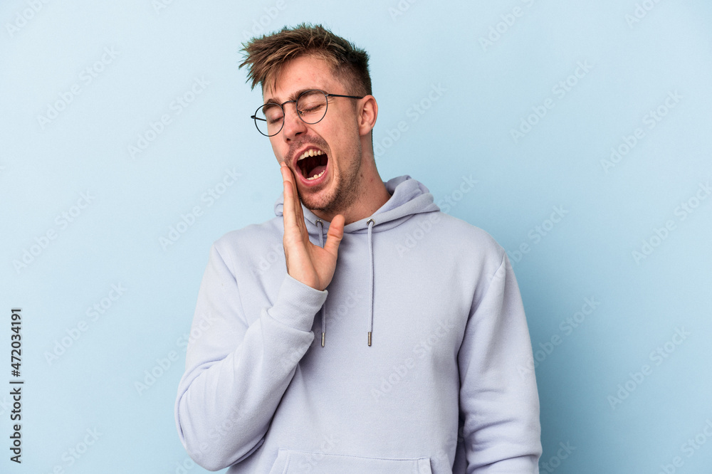 Young caucasian man isolated on blue background yawning showing a tired gesture covering mouth with hand. Young caucasian man isolated on blue background yawning showing a tired gesture covering mouth with hand.
