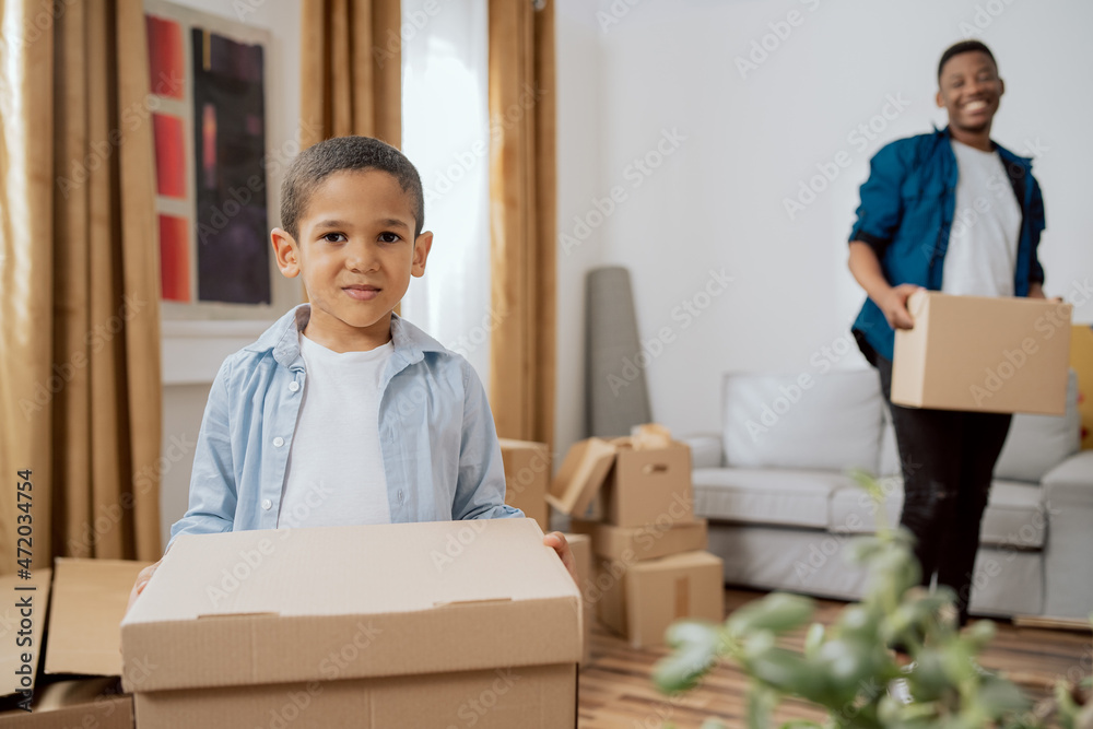 A small child bravely helps parents to carry packed things in boxes, a family moves out of the apartment to a new house, a dad proudly admires his son engaged in moving A small child bravely helps parents to carry packed things in boxes, a family moves out of the apartment to a new house, a dad proudly admires his son engaged in moving