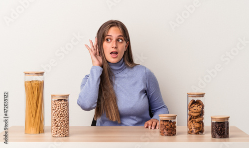 Young caucasian woman sitting at a table with food pot isolated on white background trying to listening a gossip.