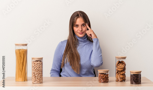 Young caucasian woman sitting at a table with food pot isolated on white background pointing temple with finger, thinking, focused on a task.