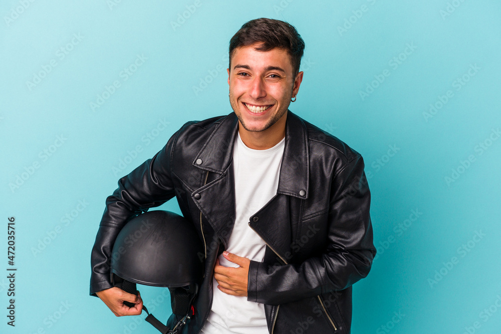 Young biker man holding helmet isolated on blue background laughing and having fun. Young biker man holding helmet isolated on blue background laughing and having fun.