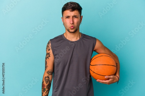 Young caucasian man playing basketball isolated on blue background shrugs shoulders and open eyes confused.