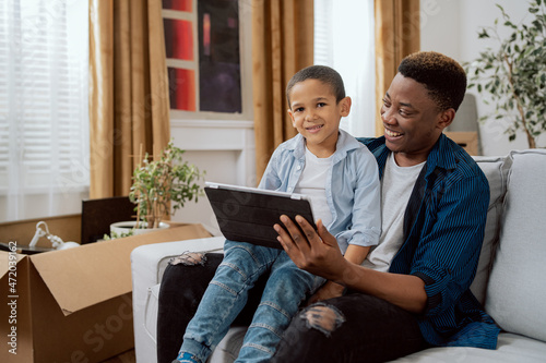 A smiling man holds a tablet in his hand, shows his son videos, funny pictures, they browse the Internet, boys laugh, father and son spend time together on the couch