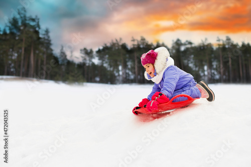childhood, leisure and season concept - happy little girl sliding down on snow saucer sled outdoors in winter over snowy forest or park background