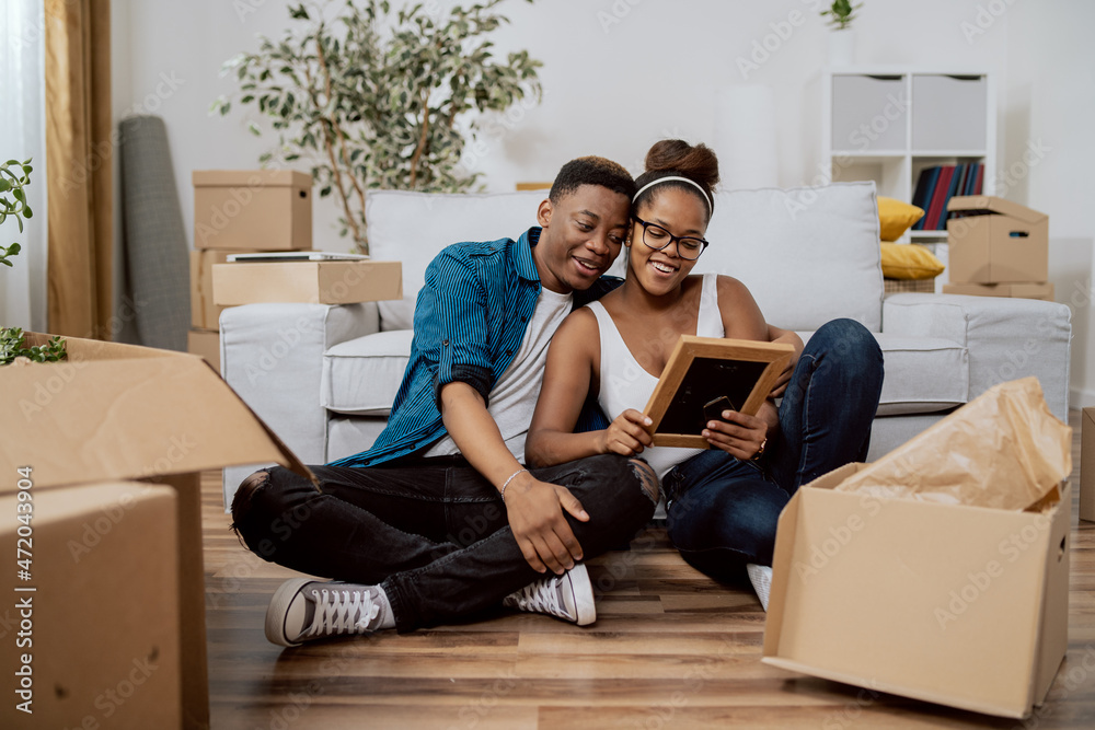 Married couple after moving to new house unpack boxes sitting on floor, woman shows husband found photo in frame from their wedding, man embraces wife, they smile reminisce Married couple after moving to new house unpack boxes sitting on floor, woman shows husband found photo in frame from their wedding, man embraces wife, they smile reminisce