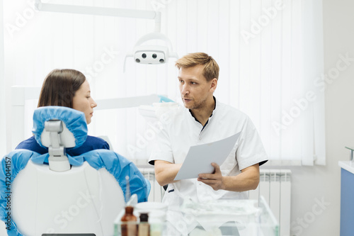 Male Professional Dentist With Gloves And Mask Holding papers photo And Show What The Treatment Will Look Like Of The Patient's Teeth. Discussion Of The Treatment Plan And Healthy Smile