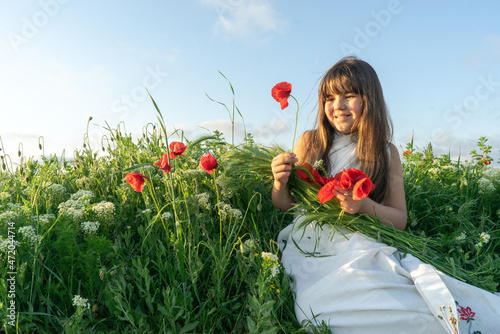 A girl in a white dress in her arms holds a bouquet of red poppies and green spike-lets of wheat, in a field of flowers with poppies and white flowers at sunset