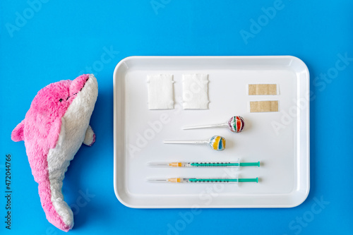 Stuffed dolphin toy by tray of vaccine injections and lollipop on blue background
