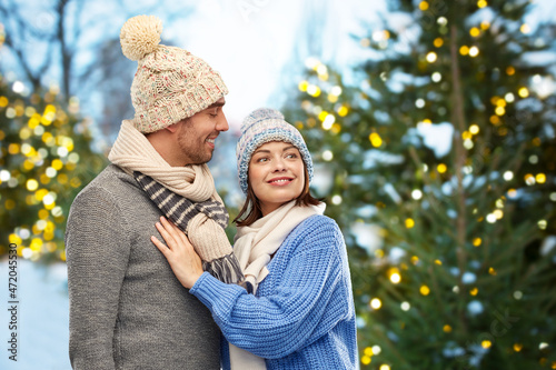 people and winter holidays concept - happy couple in knitted hats and scarves outdoors over christmas tree lights background